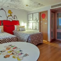 Quarto temático do hotel com personagens Snoopy na parede e roupa de cama, incluindo uma mesa redonda com desenhos.