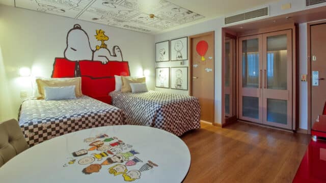 Quarto temático do hotel com personagens Snoopy na parede e roupa de cama, incluindo uma mesa redonda com desenhos.