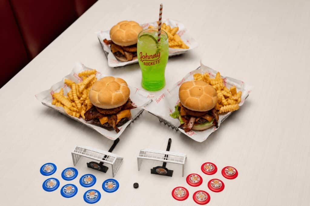 Mesa com três combos do Johnny Rockets: hambúrguer, batatas fritas e refrigerante verde, além de futebol de botão azul e vermelho.
