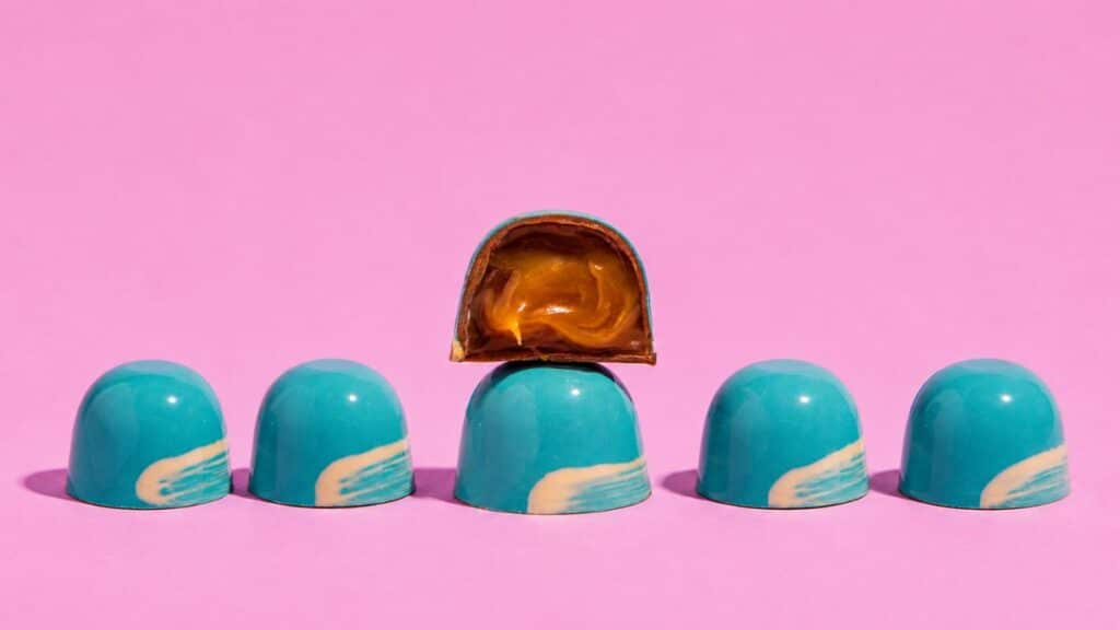 Bombons com cobertura azul e recheio caramelizado, destacados sobre fundo rosa, da Mica Chocolates no Dia Mundial do Chocolate.