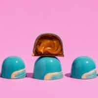 Bombons com cobertura azul e recheio caramelizado, destacados sobre fundo rosa, da Mica Chocolates no Dia Mundial do Chocolate.