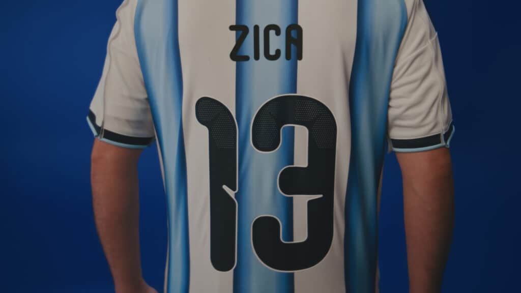 Jogador de futebol com camisa azul e branca do Agibank, com o texto “ZICA” no centro.