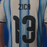 Jogador de futebol com camisa azul e branca do Agibank, com o texto “ZICA” no centro.