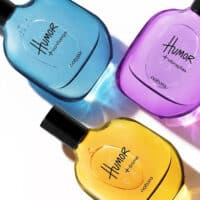 Três frascos da linha Humor da Natura nas cores azul, roxa e amarela, com fragrâncias e rótulos visíveis