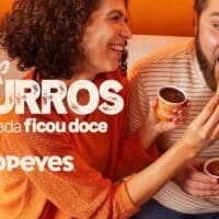 Casal saboreando Pop Churros do Popeyes, com o produto em destaque e um copo de molho ao lado.