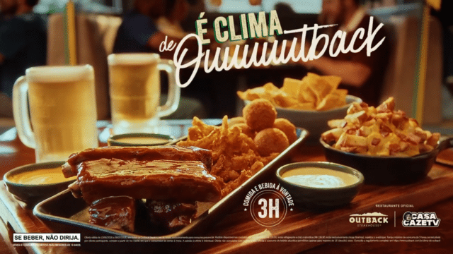 Mesa com porções de comida do Outback, incluindo costela, nuggets e molho, ao lado de canecas de cerveja