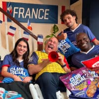 Grupo em festa com garrafa Smirnoff Ice e pacotes do Club Social Snack, ao lado de bandeiras e cartaz “FRANCE”.