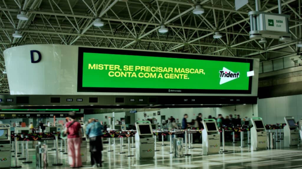 Placa publicitária da Trident no Aeroporto Internacional Tom Jobim, com texto verde e fundo no saguão.