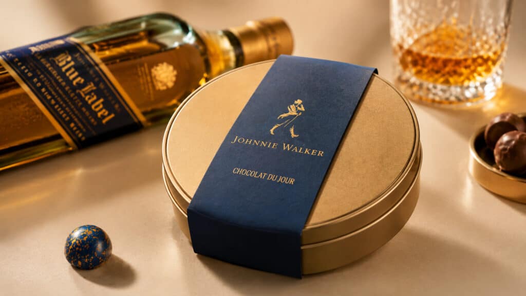 Embalagem azul “Chocolat du Jour” da Johnnie Walker sobre mesa com garrafa e taça ao fundo, em pop-up store