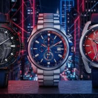 Três relógios Citizen temáticos do Homem-Aranha com mostradores azul, vermelho e metálico, em cenário futurista urbano