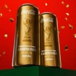 Duas latas douradas da Coca-Cola com a frase “Campeones España” em fundo vermelho com confetes.