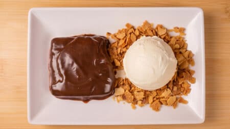Torta Al Cioccolato com cobertura de chocolate e sorvete, servida em prato branco sobre mesa de madeira.