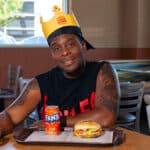 Homem em restaurante do Burger King com chapéu, segurando e ao lado de uma Fanta, com sanduíche em uma mesa
