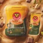 Embalagem de Cappuccino Chocolate com Pistache da 3 Corações, com pistaches e pedaços de chocolate ao fundo.