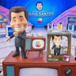 Boneco colecionável do Silvio Santos em uma TV, na bancada do estúdio com o logo do SBT ao fundo.