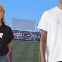 Modelo veste camiseta branca e preta com logo da Levi’s coberto, diante de estádio de futebol em dia de partida da Copa do Mundo.