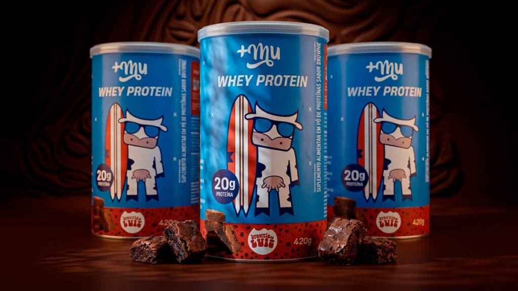 Whey protein Mais Mu e Brownie do Luiz sabor brownie Latas de whey protein da Mais Mu e do Brownie do Luiz, sabor brownie, com cubos e pedaços de brownie ao fundo.