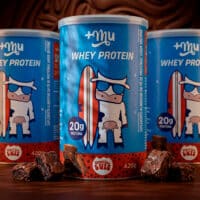 Latas de whey protein da Mais Mu e do Brownie do Luiz, sabor brownie, com cubos e pedaços de brownie ao fundo.
