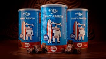 Whey protein Mais Mu e Brownie do Luiz sabor brownie Latas de whey protein da Mais Mu e do Brownie do Luiz, sabor brownie, com cubos e pedaços de brownie ao fundo.