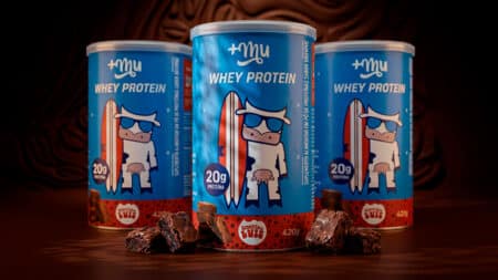 Whey protein Mais Mu e Brownie do Luiz sabor brownie Latas de whey protein da Mais Mu e do Brownie do Luiz, sabor brownie, com cubos e pedaços de brownie ao fundo.