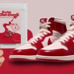 Tênis Air Jordan 1 High OG vermelho e branco com charms de unhas e mãos femininas exibindo esmalte vermelho.