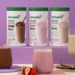 Bebidas proteicas Herbalife em potes e copos com mãos segurando um líquido, em fundo roxo e rosa.
