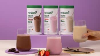 Bebida proteica Herbalife em embalagens e copos Bebidas proteicas Herbalife em potes e copos com mãos segurando um líquido, em fundo roxo e rosa.