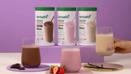 Bebidas proteicas Herbalife em potes e copos com mãos segurando um líquido, em fundo roxo e rosa.