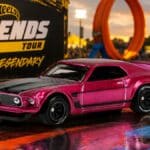 Carro Hot Wheels Legends Tour rosa em cenário de evento, com banners “Hot Wheels Legends” e “Be Legendary” ao fundo.