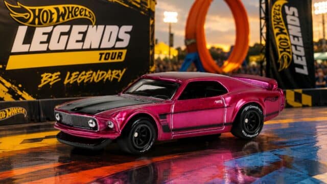 Carro Hot Wheels Legends Tour rosa em cenário de evento, com banners “Hot Wheels Legends” e “Be Legendary” ao fundo.