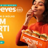 Campanha do Popeyes Minis com mulher provando sanduíche em versão mini. Texto: “Chegaram os novos” e “Popeyes Minis”.
