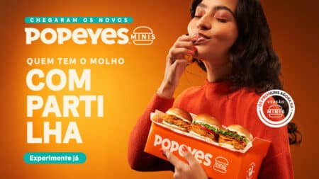 Popeyes Minis: promoção com sanduíches em miniatura Campanha do Popeyes Minis com mulher provando sanduíche em versão mini. Texto: “Chegaram os novos” e “Popeyes Minis”.