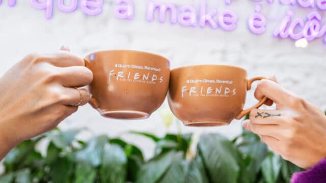 Duas mãos brindando com canecas marrons com a marca “FRIENDS”.