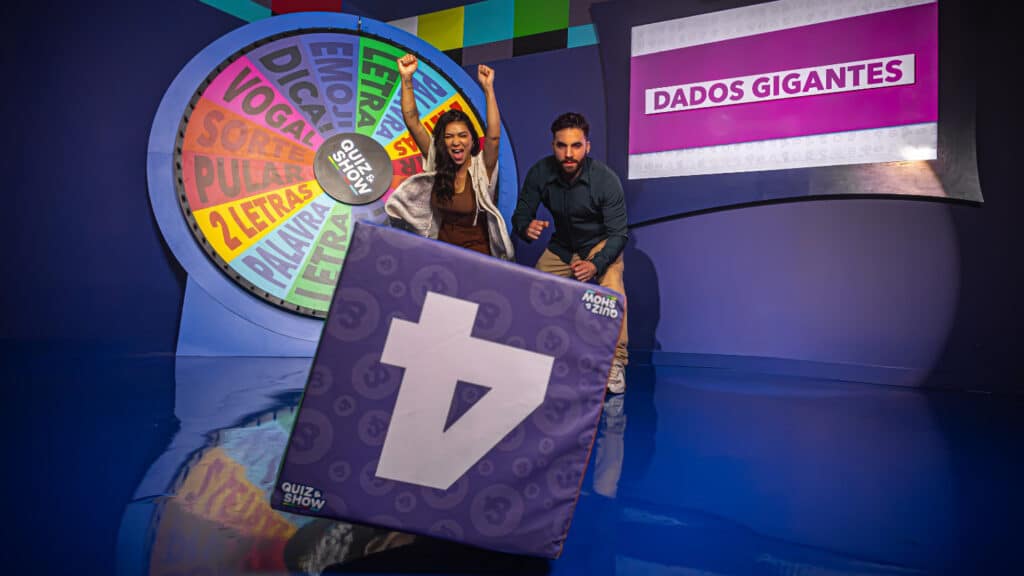 Apresentadores posam em cenário do Quiz&Show, com painel roxo e placa grande de seta no palco