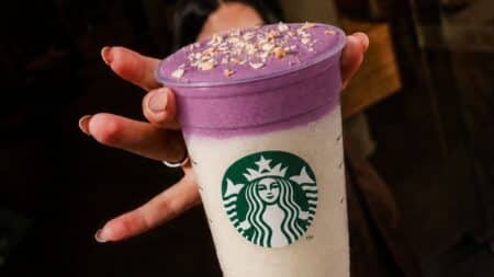 Starbucks Brasil lança bebida de ube roxa Copo da Starbucks com bebida roxa de ube, com tampa e chantilly, segurado por mãos em ambiente interno.