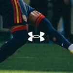 Chuteira de futebol em ação durante partida, com o logo Under Armour no calçado e número 106 na imagem.
