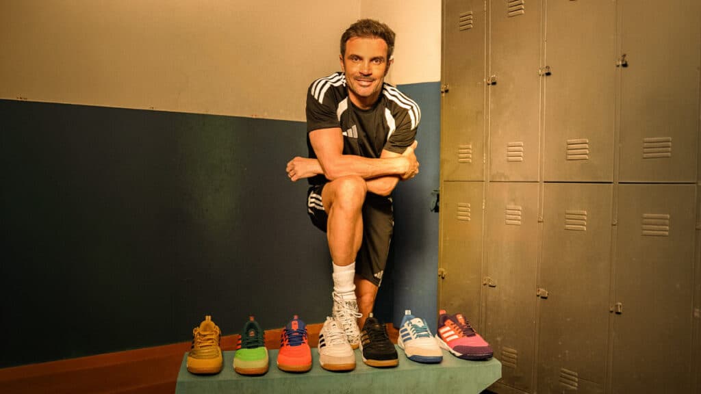 Homem com uniforme da Adidas posando ao lado de vários tênis de futsal, em ambiente interno com armários