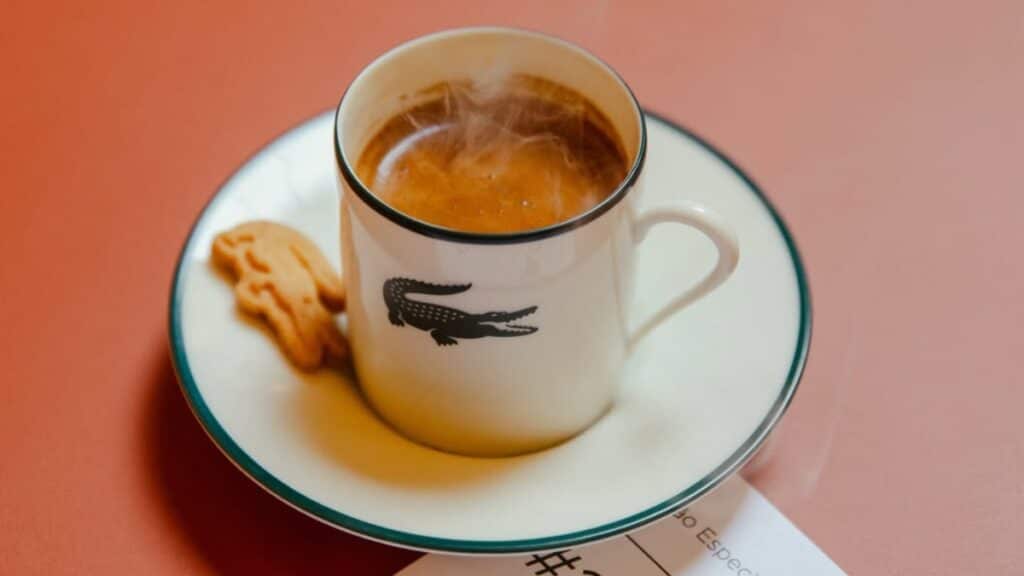Xícara de café com logo da Lacoste em pires, sobre fundo rosado, sugerindo o Café Lacoste no Brasil.