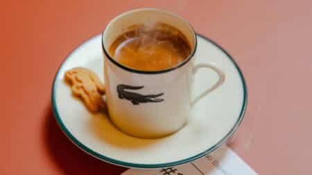 Café Lacoste no Brasil: xícara com logo da Lacoste Xícara de café com logo da Lacoste em pires, sobre fundo rosado, sugerindo o Café Lacoste no Brasil.