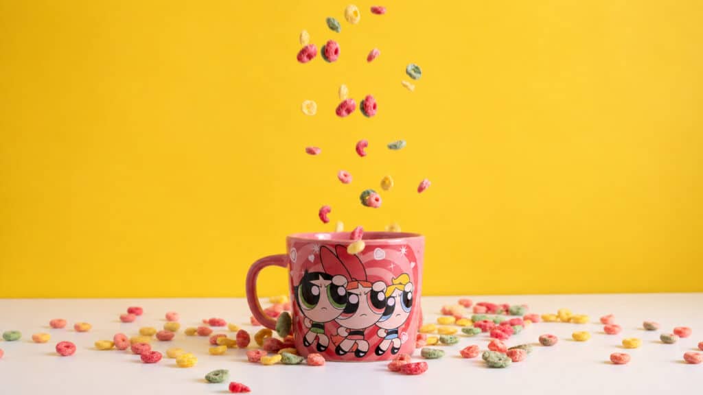 Caneca colecionável do Cartoon Network com personagens, sobre mesa com confetes coloridos em fundo amarelo.