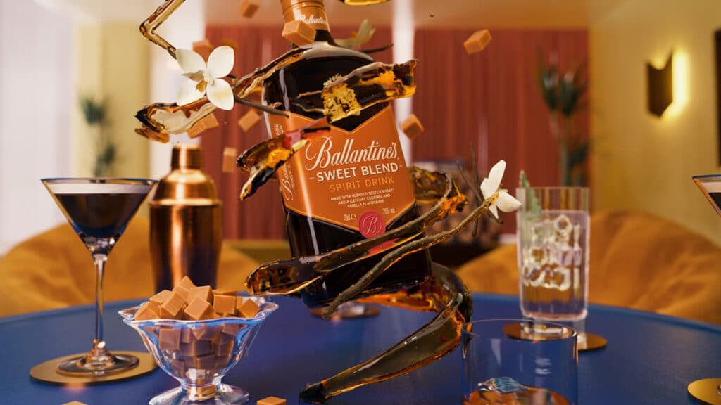 Frente de uma garrafa Ballantine's Sweet Blend em mesa de degustação, com copos e doces ao fundo.