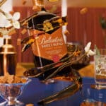 Frente de uma garrafa Ballantine's Sweet Blend em mesa de degustação, com copos e doces ao fundo.
