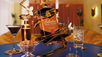 Frente de uma garrafa Ballantine's Sweet Blend em mesa de degustação, com copos e doces ao fundo.