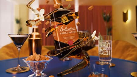 Frente de uma garrafa Ballantine's Sweet Blend em mesa de degustação, com copos e doces ao fundo.