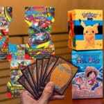 Coleção de cards e caixas de brinquedos Pokémon, Megaevolução, Harry Potter e Naruto sobre mesa, com mão segurando cartas.