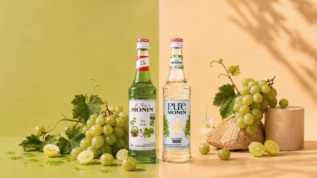 Garrafa de xarope Monin sabor uva verde e uvas ao lado, em cenário com fundo claro