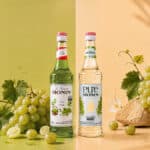 Garrafa de xarope Monin sabor uva verde e uvas ao lado, em cenário com fundo claro