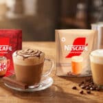 Embalagens de Nescafé Cappuccino em sachê Tradicional e KitKat, com copos de bebida cremosa sobre a mesa