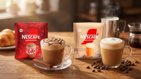 Nescafé Cappuccino Tradicional e KitKat em sachê Embalagens de Nescafé Cappuccino em sachê Tradicional e KitKat, com copos de bebida cremosa sobre a mesa