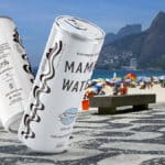 Lata de água Mamba-rio em destaque na orla do Rio de Janeiro, com quiosques e montanhas ao fundo.